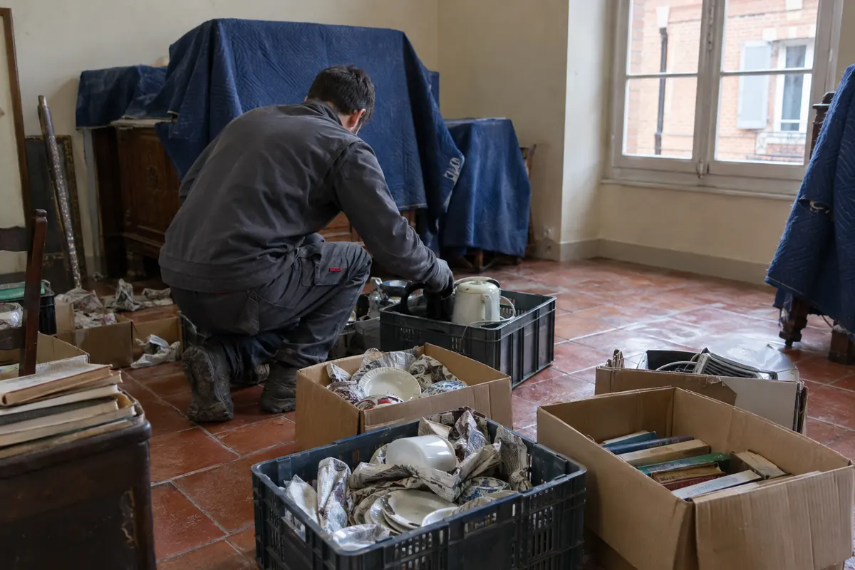 Intervenant triant vaisselle, livres et petit électroménager dans des caisses, dans une pièce de maison en cours de débarras